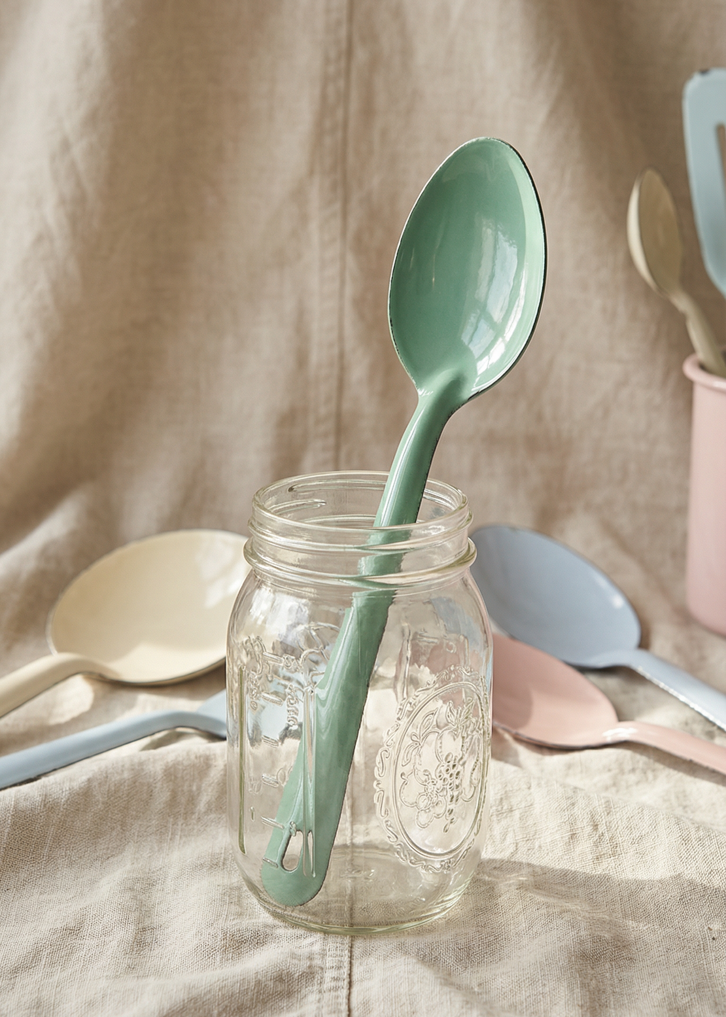 Green spoon in a glass jar with other kitchen utensils on a beige fabric background