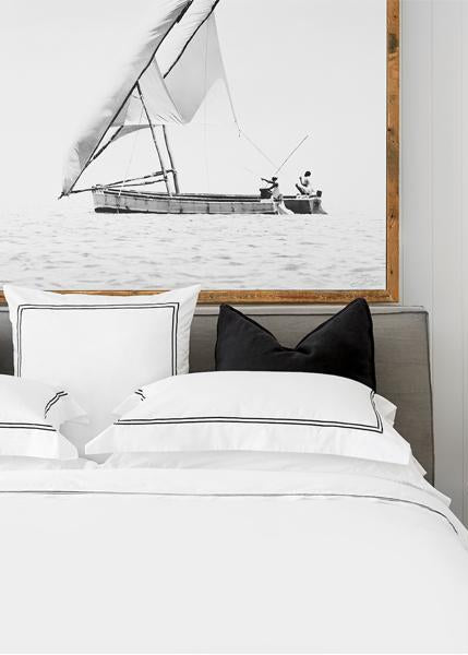 A made bed with white bedding, featuring a flat sheet and pillowcases, in a room with a sailing-themed artwork on the wall and a decorative plant on the side table.