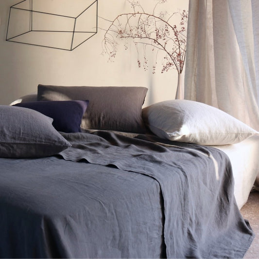 A linen flat top sheet with stone blue piping is displayed on a bed, showcasing its soft texture and the contrast piping detail.