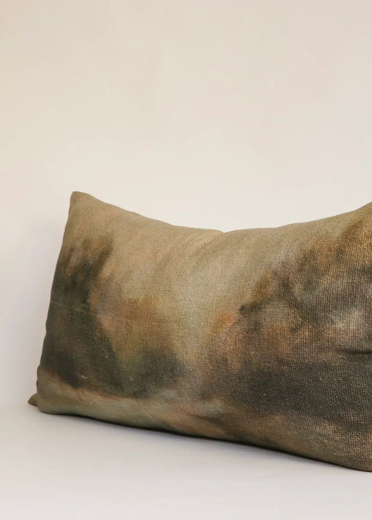 A rectangular lumbar throw pillow with a textured, abstract hand-dyed pattern in earthy tones of brown, olive green, and beige, shown against a plain off-white background.