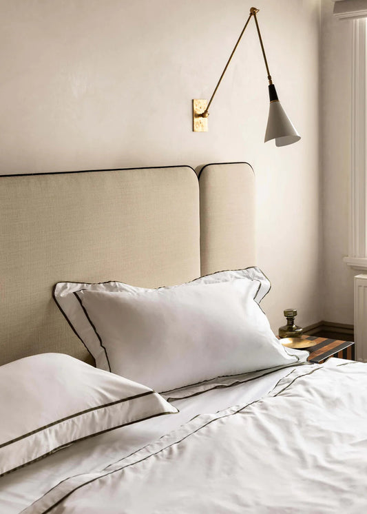 A pair of white pillowcases with navy grosgrain ribbon trim, displayed on a bed with a beige headboard.