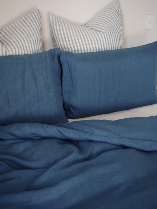 A blue linen euro pillowcase displayed on a bed with other bedding items in the background.