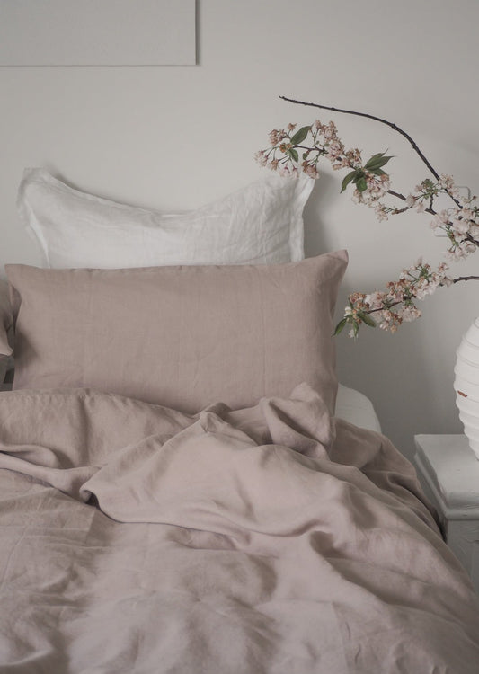 Neatly made bed with beige bedding and white pillows, featuring a branch with flowers on a light gray wall.