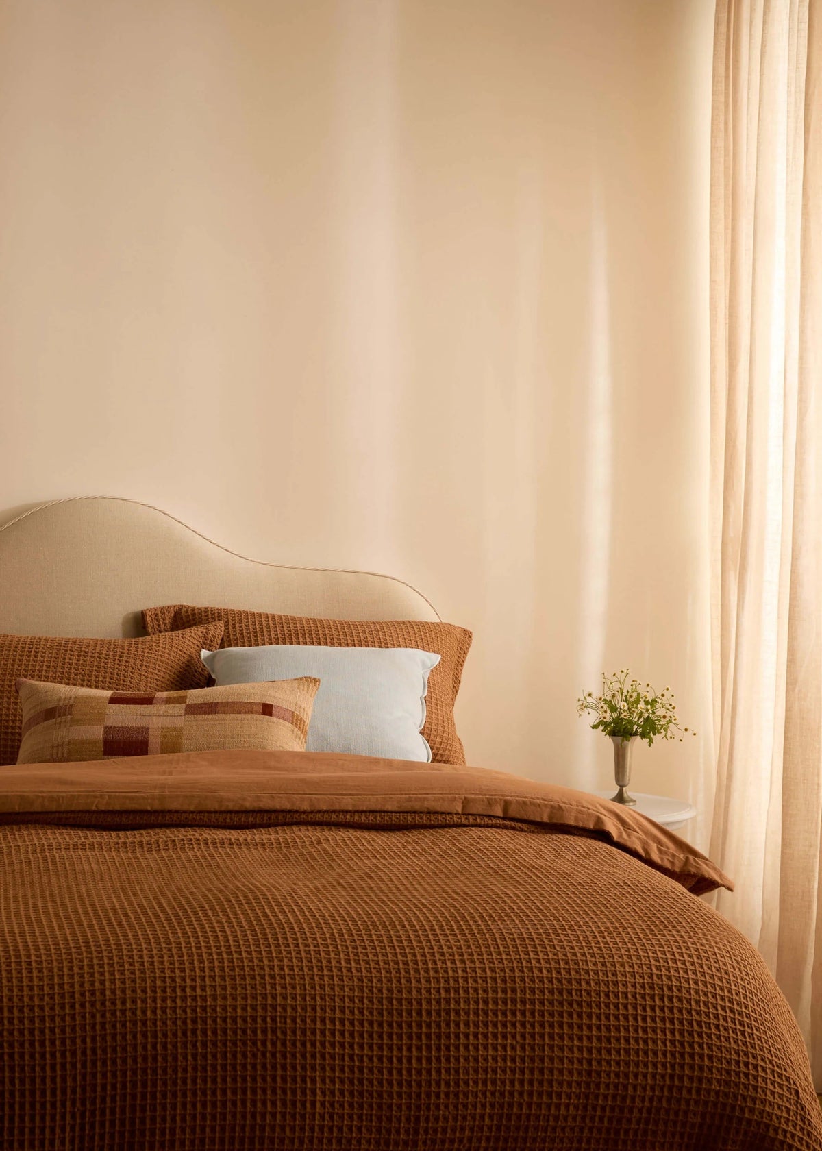 A cozy bedroom scene with a bed dressed in warm brown textured bedding, matching pillows, a cream upholstered headboard, sheer curtains, and a small vase of flowers on a bedside table.