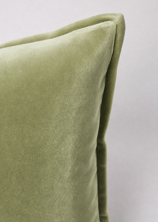 A close-up of a sage green velvet throw pillow corner, showing the soft texture and piped edge against a neutral light gray background.