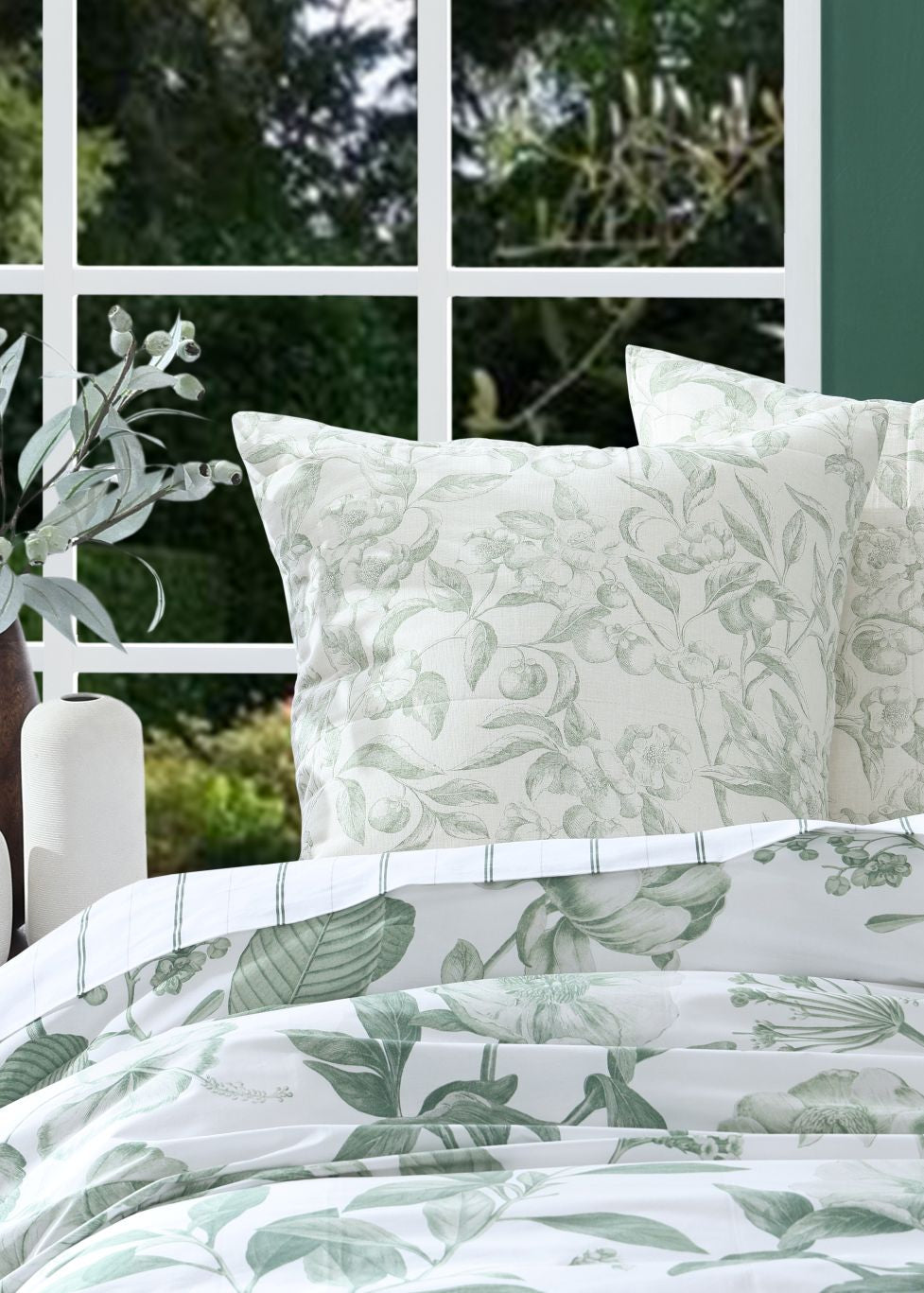 Floral-patterned bedding set with matching pillows on a bed, against a green wall and window.