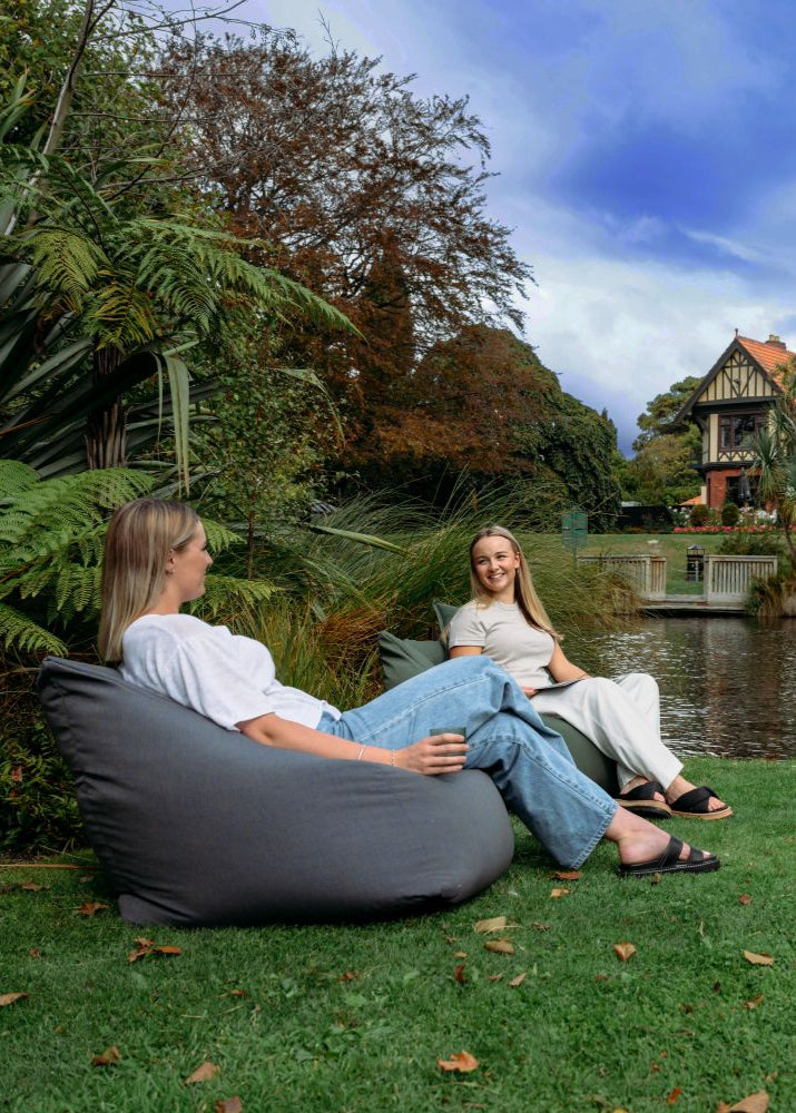 Two people sitting on a bean bag by a pond with a house and trees in the background