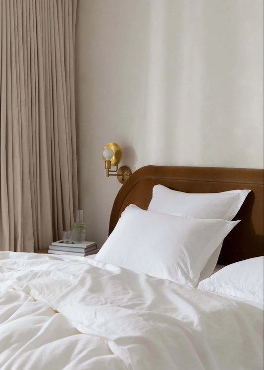 A single white pillowcase on a bed, with a wooden headboard and a wall sconce in the background, curtains to the side.