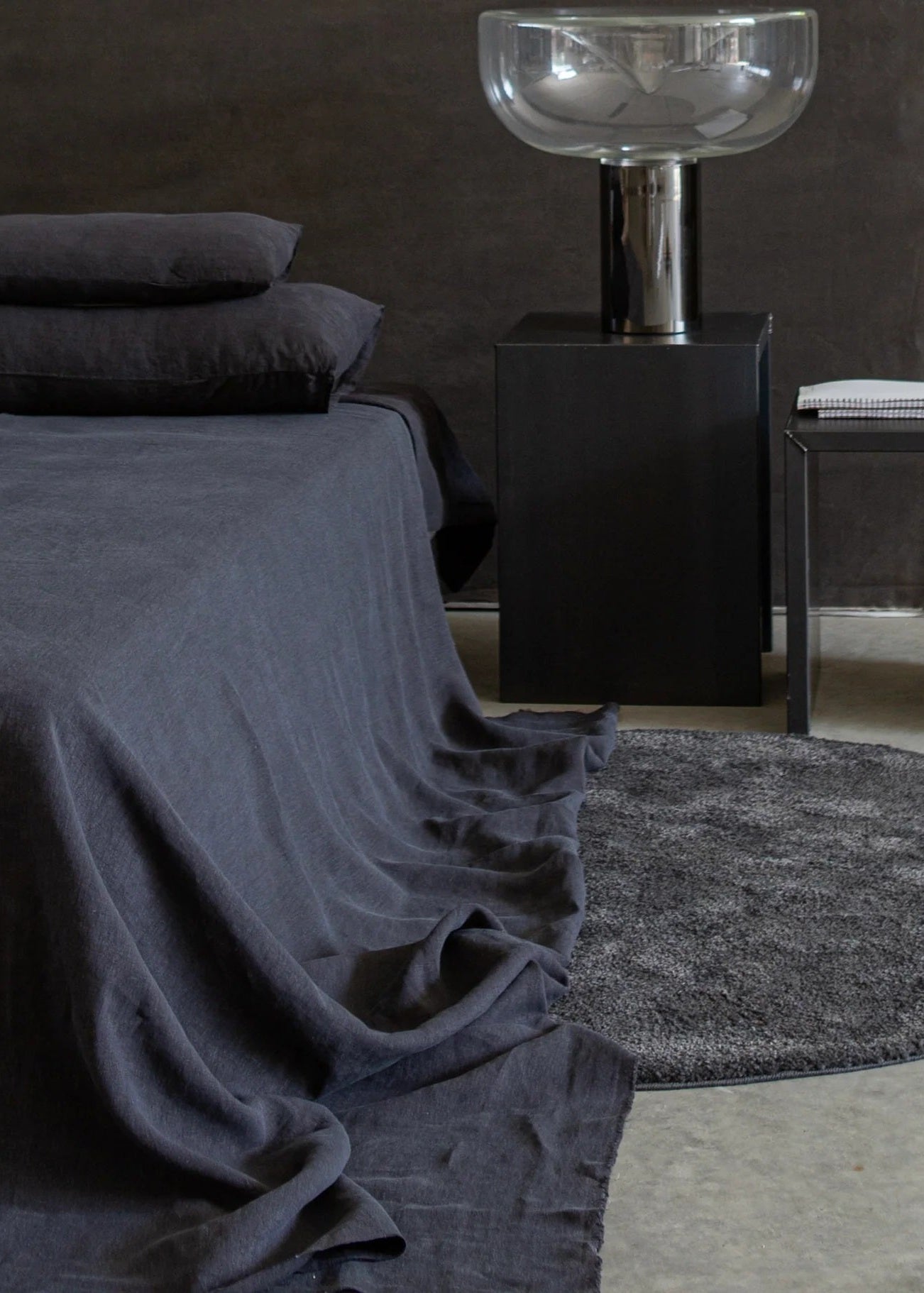 A black linen bed cover with piping detail, displayed on a bed in a dark room setting with pillows, a nightstand, and a floor lamp.