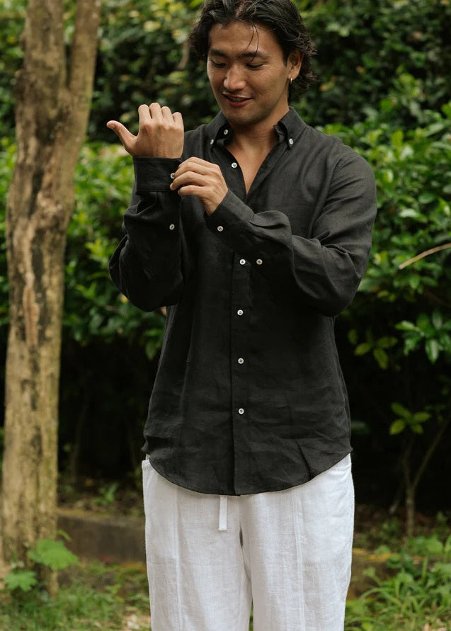 Man wearing a black shirt and white pants standing outdoors with greenery in the background
