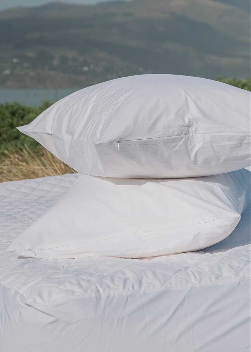 Two white pillow protectors on a bed, with a zippered pillow on top and a flat pillow on the bottom, against a background of a natural landscape.