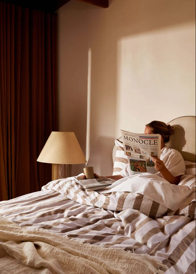 A person lying on a bed reading a newspaper, with a striped duvet cover in a color that complements the bedding.