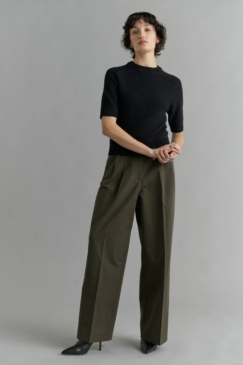 Woman wearing a black top and olive green pants against a gray background