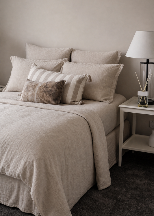 A neatly made bed with layered beige and cream linens, including striped and textured pillows, set in a minimalist bedroom with a white nightstand and lamp.