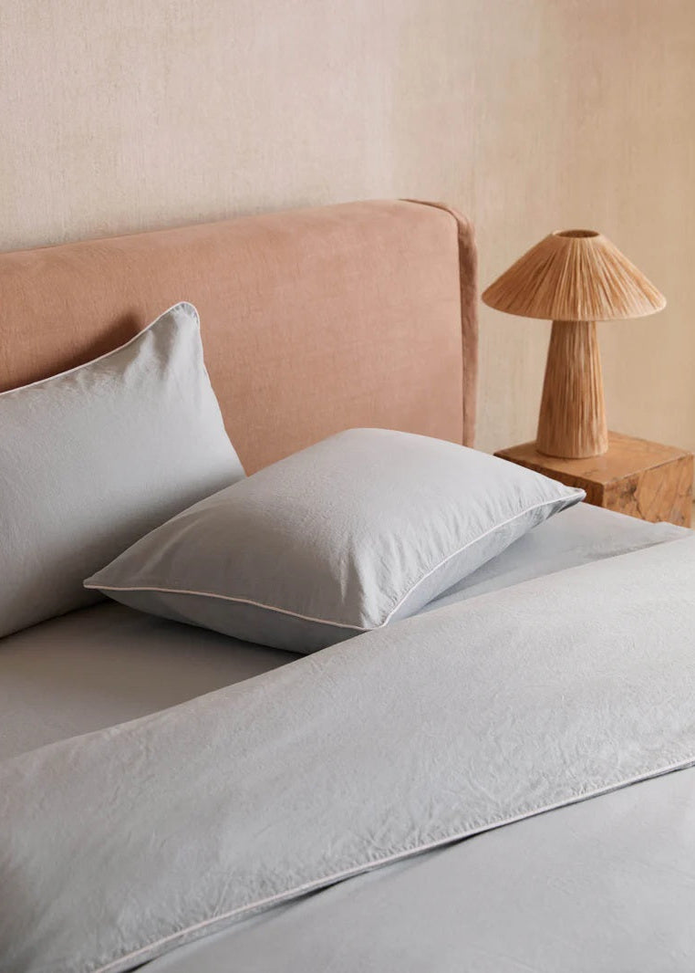 Neatly made bed with light gray bedding and pillows, wooden nightstand with lamp.
