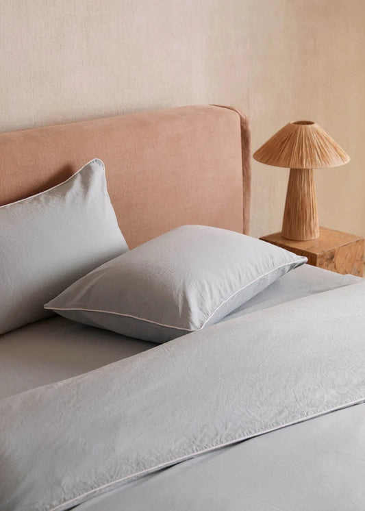 Neatly made bed with light gray bedding and pillows, wooden nightstand with lamp.