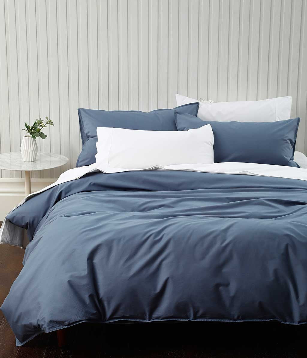 A denim blue duvet cover set on a bed, with two white pillows and a smaller blue pillow, in a room with a light-colored wall and a small plant beside the bed.