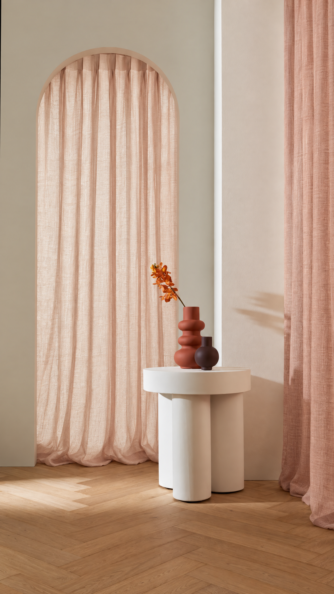 A styled interior scene with blush pink curtains hanging in an arched window opening and along the right wall, set in a room with light wood flooring and a small white side table holding a terracotta vase with orange flowers.