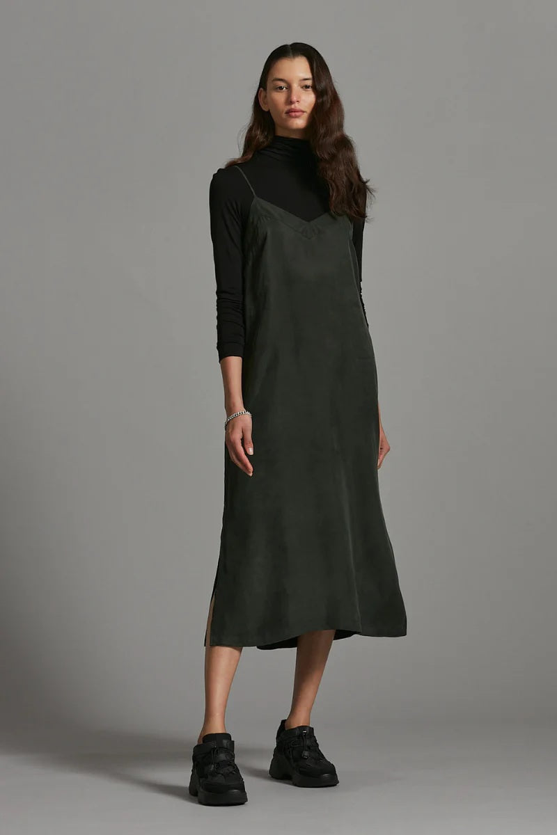 Woman wearing a dark green slip dress over a black top against a gray background