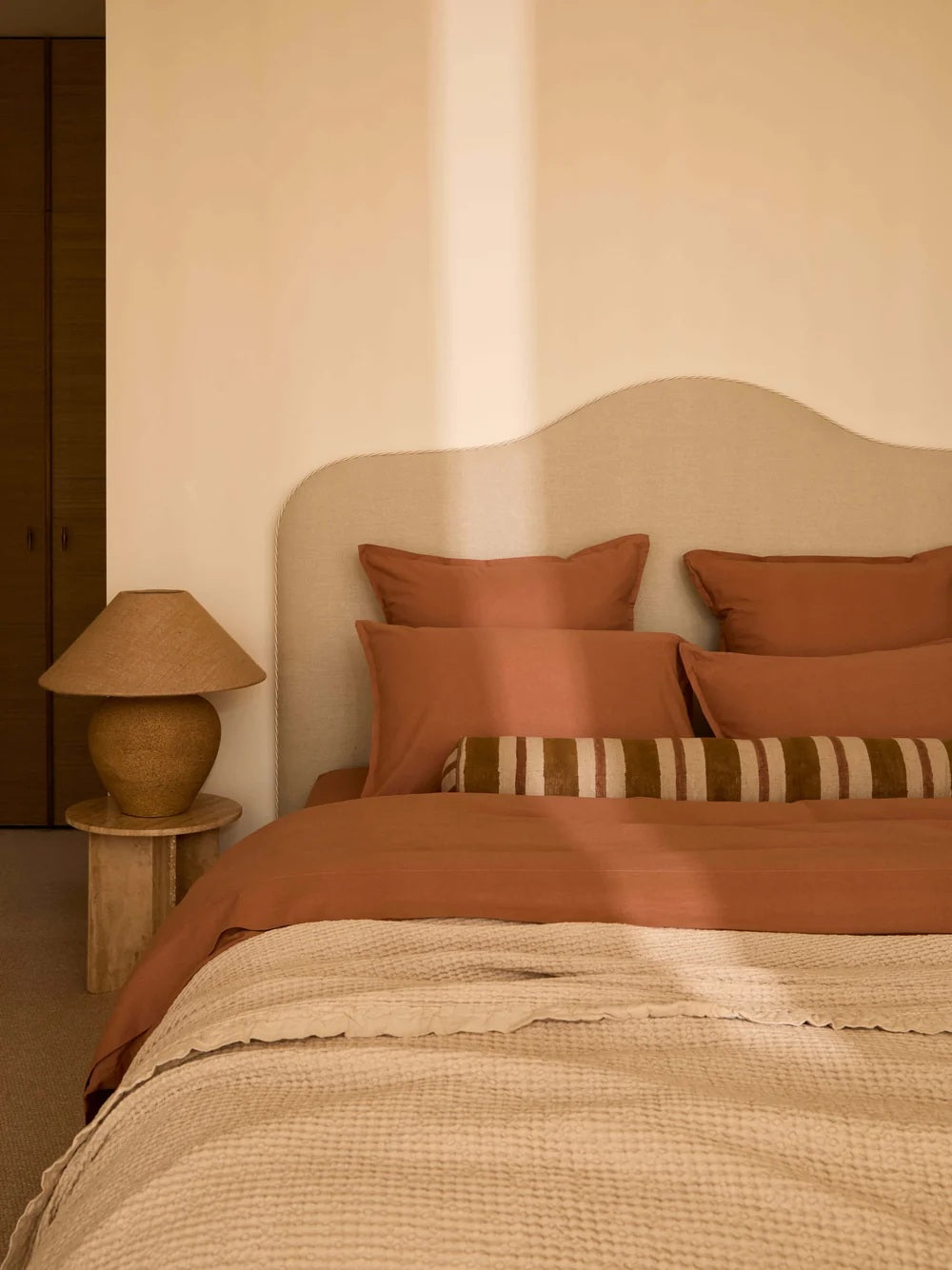 A cozy bedroom scene with a bed dressed in rust-orange bedding, matching pillows, a cream upholstered headboard, a striped lumbar pillow, and a warm-toned table lamp on a side table.