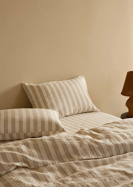 A neatly made bed with beige and cream striped bedding, including two pillows and a textured duvet, set against a warm beige wall with a dark wood table lamp on the right.