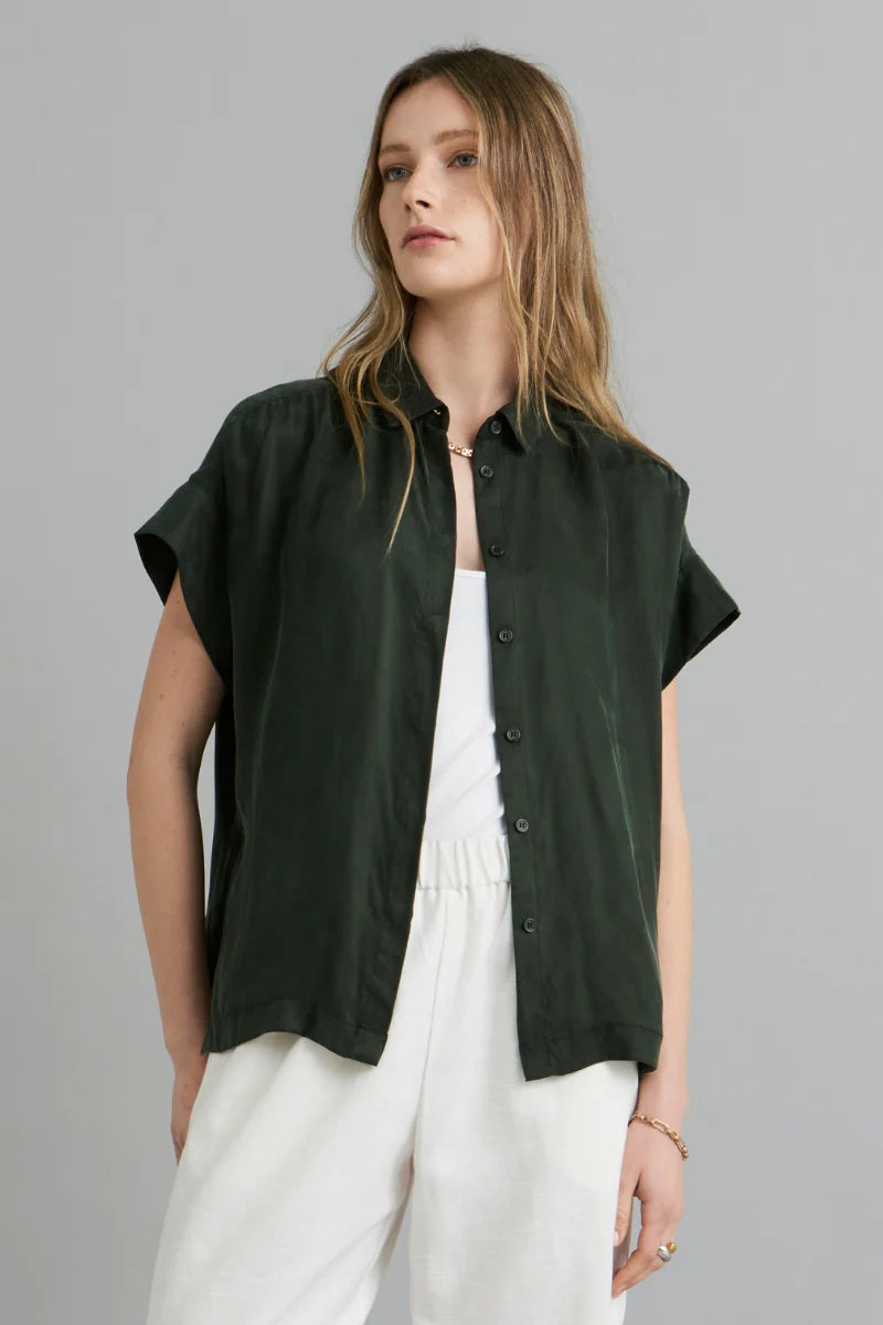 Woman wearing a dark green sleeveless blouse over a white top with light-colored pants on a gray background