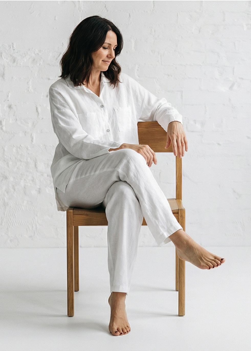 Woman sitting on a wooden chair wearing a white outfit against a white background
