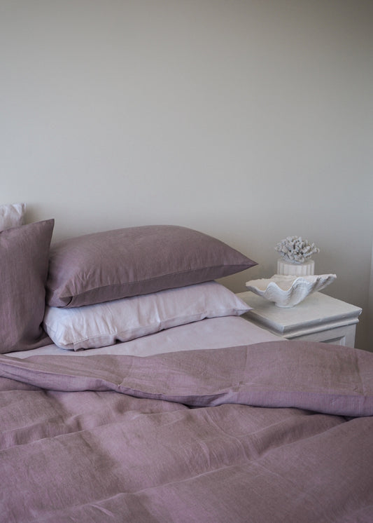 A pair of pink linen pillowcases on a bed, with a pillow, a book, and a decorative object on the side table.