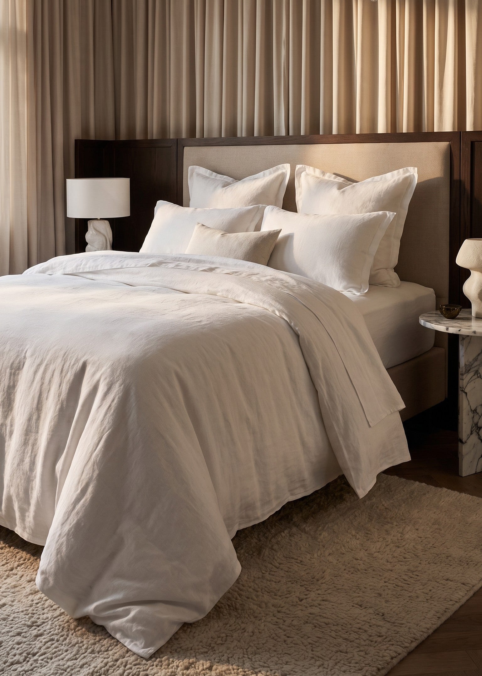 A neatly made bed with white and cream bedding in a luxurious bedroom setting, featuring a dark wood headboard, beige curtains, a crystal chandelier, and a plush rug.