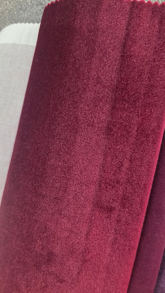 A close-up of a hand holding a rolled piece of plush velvet fabric in deep burgundy, with other fabric swatches partially visible behind it.
