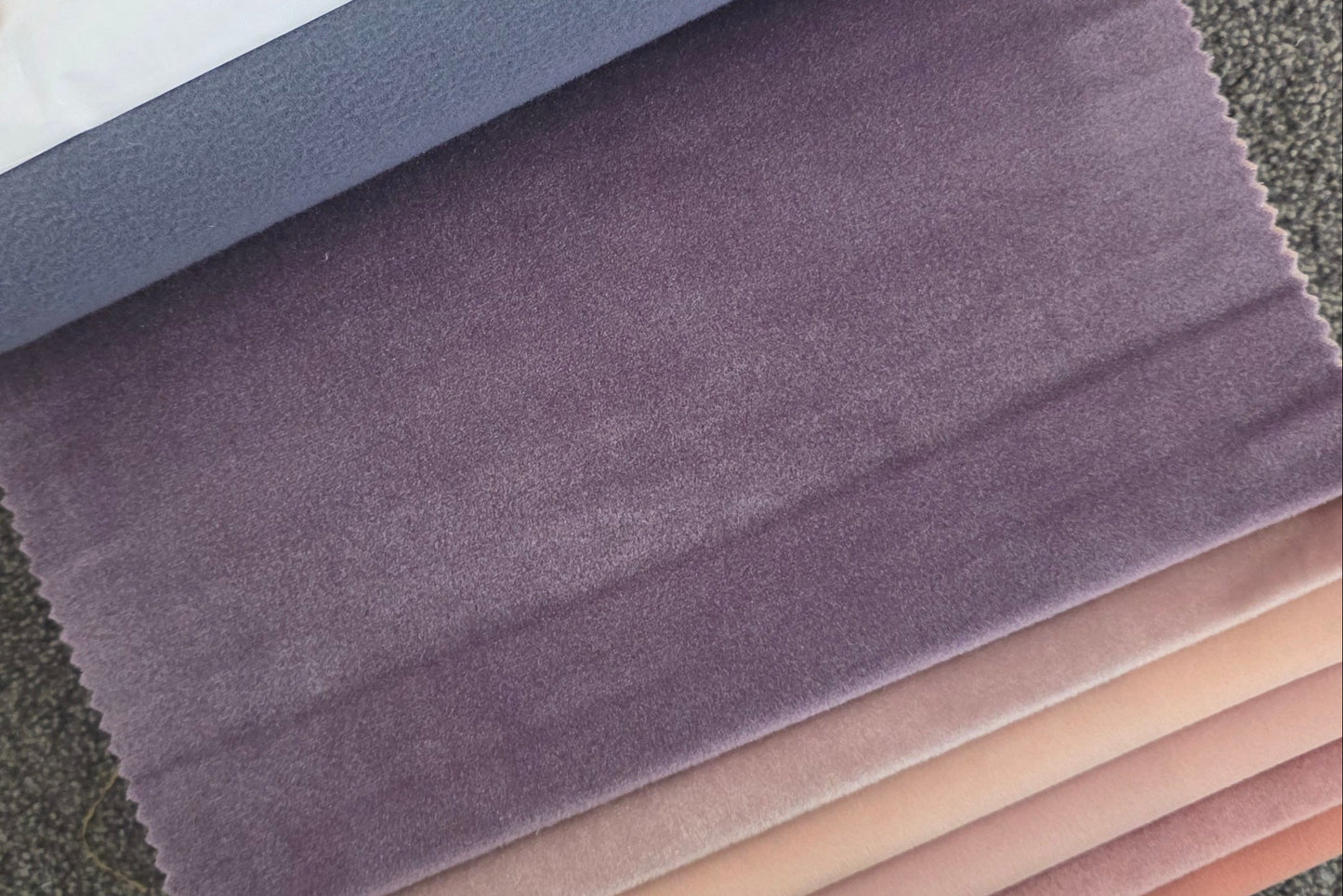 A stack of velvet fabric swatches in various colors, including purple, pink, orange, and yellow, with a hand holding the top purple swatch. A white label on the top swatch reads 'Velvet' and 'W'.