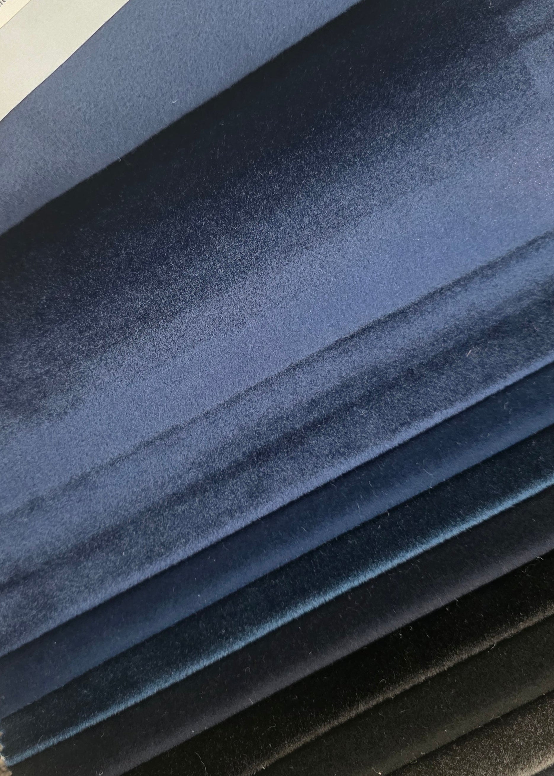 A close-up of a fabric swatch book showing layered velvet material samples in shades of blue, navy, gray, and black. A white paper label at the top reads 'Wondra Twilight'.