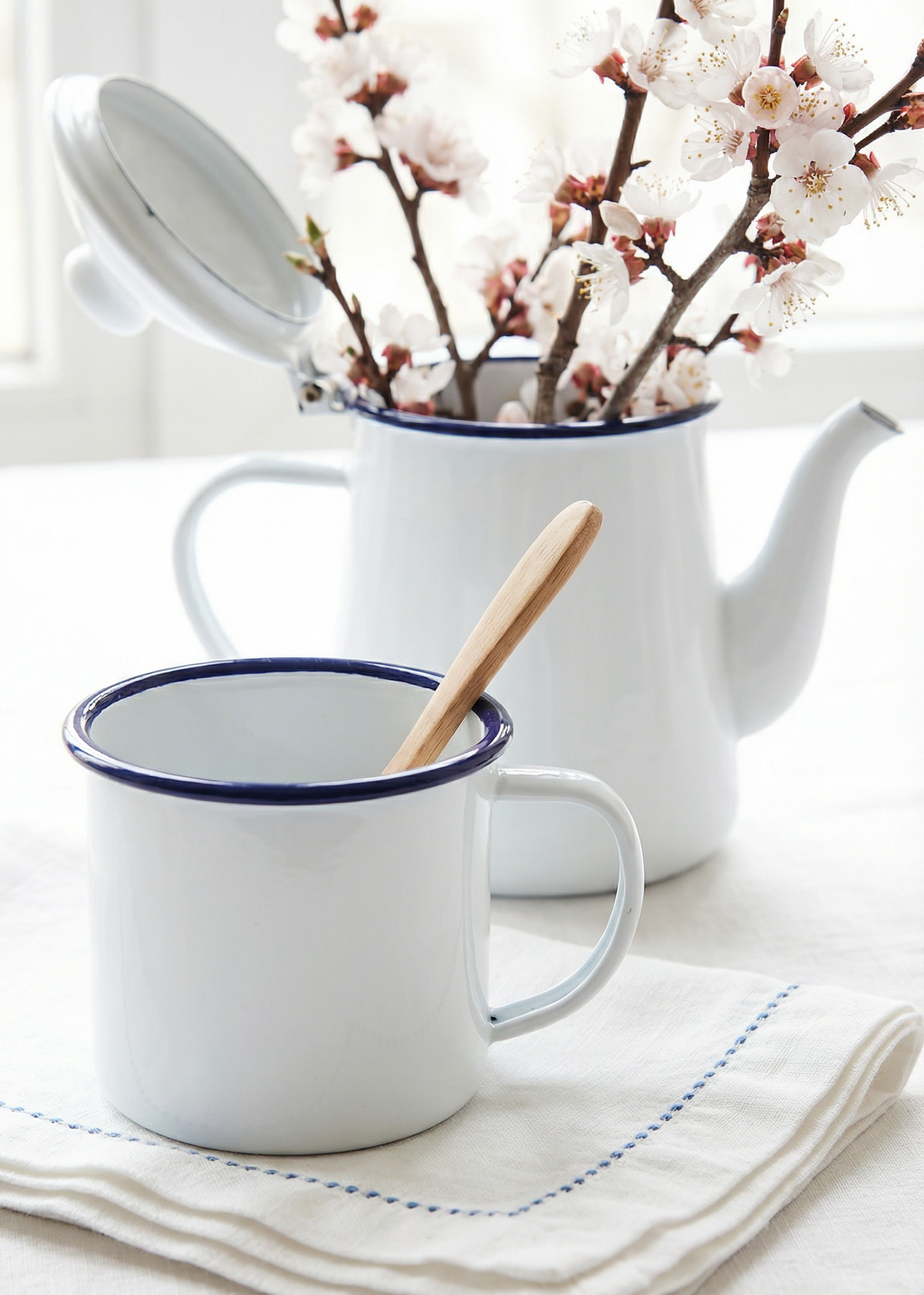 White teapot with blue rim and a white mug with blue rim on a light surface, accompanied by cherry blossom branches.