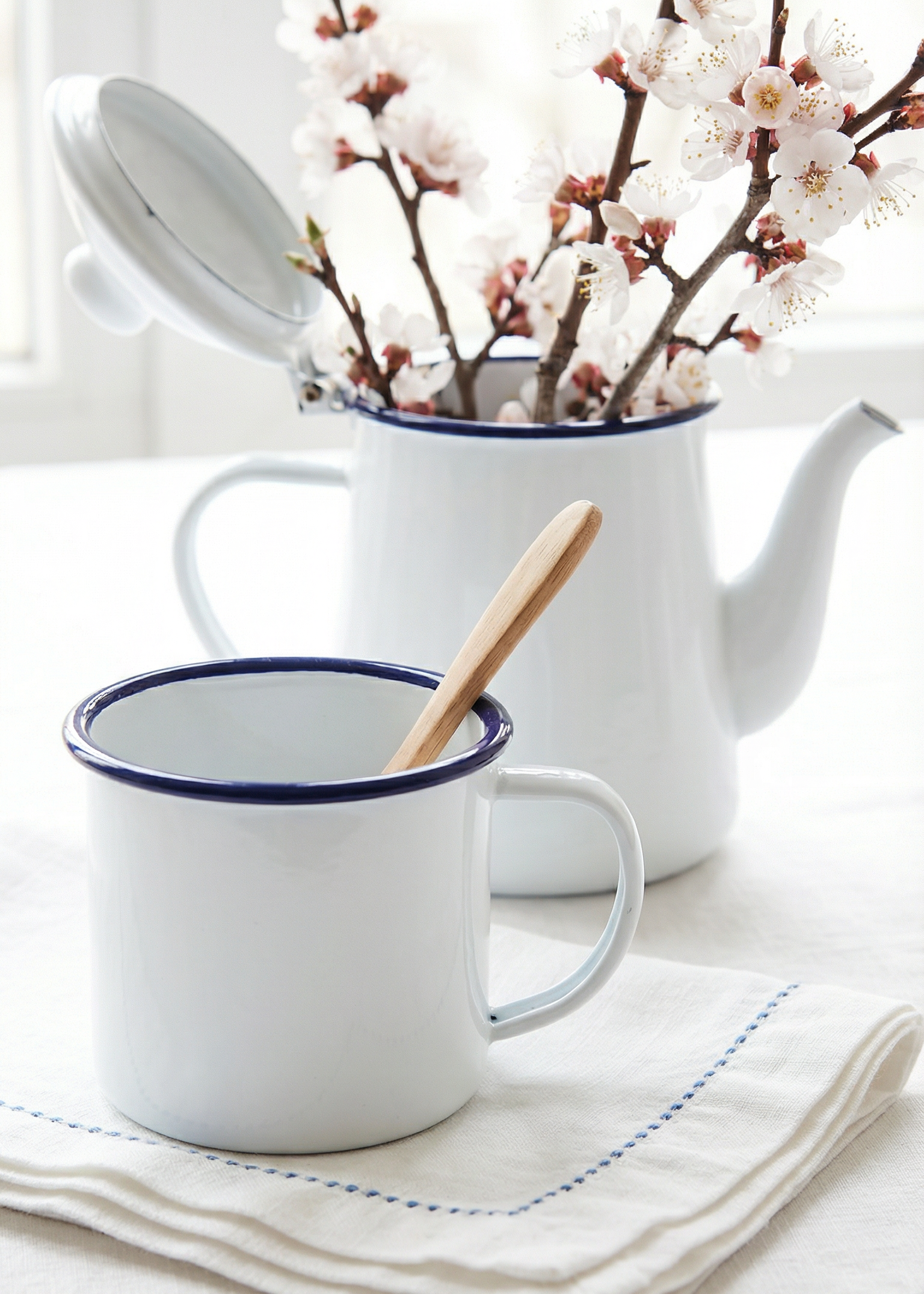 White teapot with blue rim and a white mug with blue rim on a light surface, accompanied by cherry blossom branches.