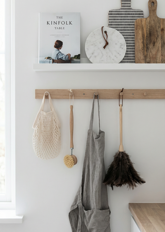 Kitchen wall with hooks holding apron, broom, and cleaning tools with a book titled 'The Kinfolk Guide' on a shelf.