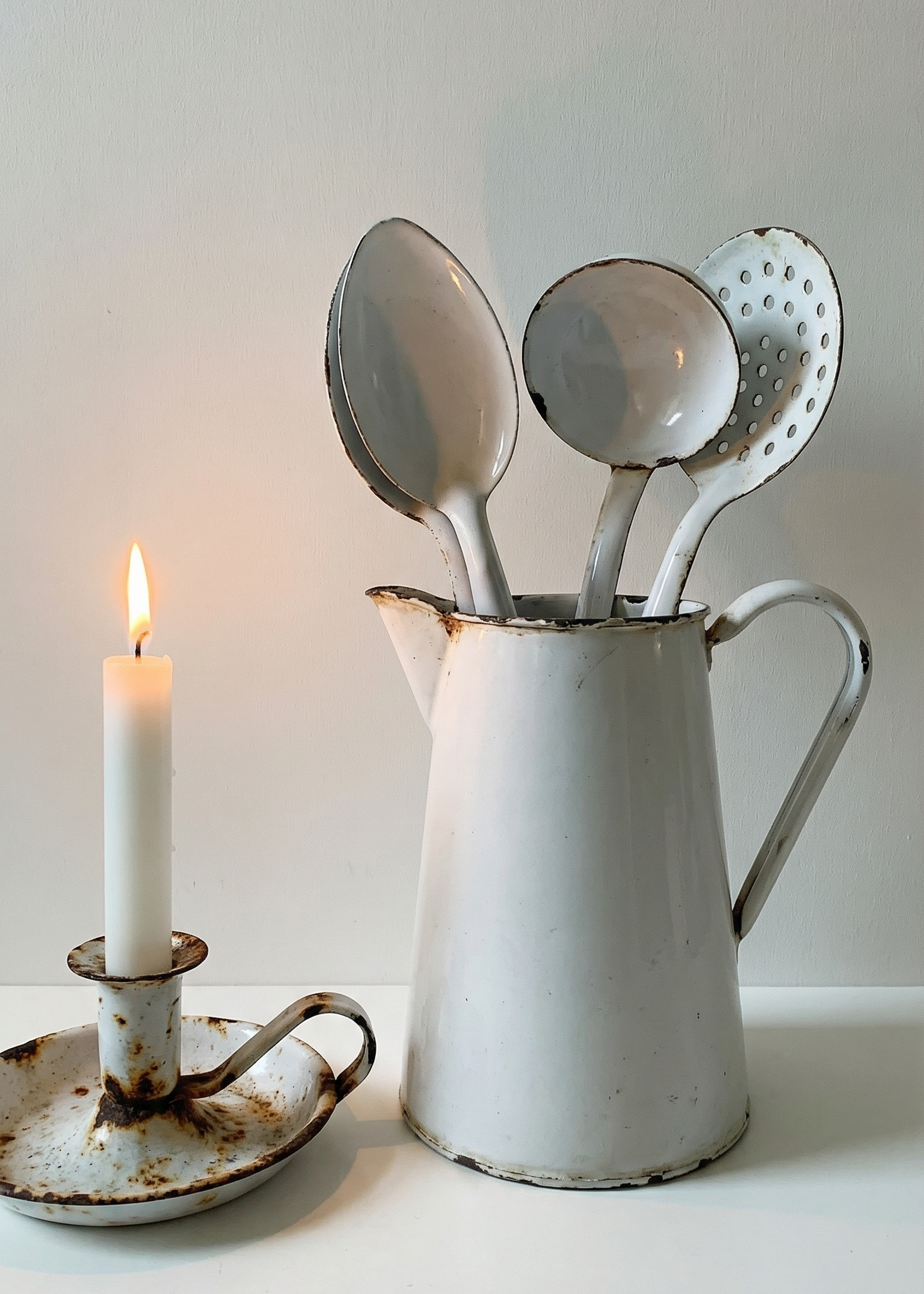 Vintage white pitcher with utensils next to a candle on a white surface