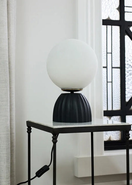 Table lamp with a white globe shade and black base on a small table next to a window.