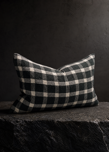 A rectangular throw pillow with a black and off-white buffalo plaid pattern, resting on a dark textured stone surface against a dark gray background.