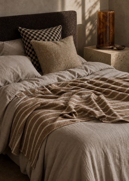 A cozy bed styled in a warm, minimalist bedroom with textured beige walls and dramatic natural light casting leaf shadows. The bed features neutral beige and brown striped bedding, a dark brown textured headboard, a black-and-beige checkered accent pillow, and a rustic amber glass lamp on a matching stone side table.