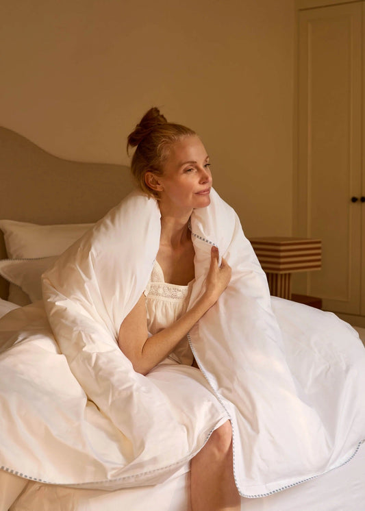A woman sits on a bed wrapped in a large white duvet or comforter in a softly lit bedroom. The bedding is voluminous and plush, with the woman looking off-camera.