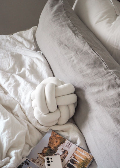 A white pure linen body pillow with a subtle textured pattern, placed on a grey sofa, with a book and a grey pillow on the sofa as well.