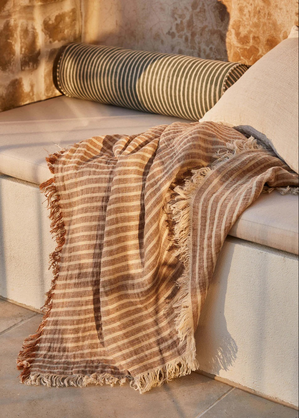A toffee-colored, striped pure linen throw blanket draped over the arm of a white sofa, with a cushion and part of a striped rug visible in the background.