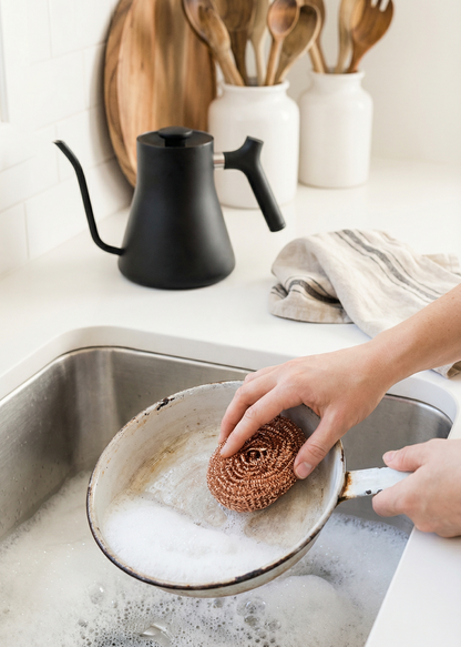 Person washing a pot with a scrubber in a kitchen sink, with a black kettle and wooden utensils in the background.