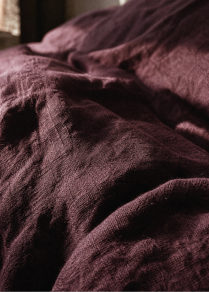 A close-up of a dark plum purple textured fabric, likely a linen throw blanket or bedspread, softly draped in the light of a window.
