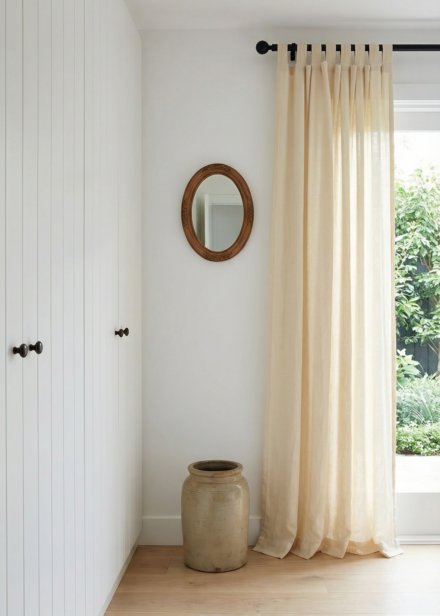 Nest living room with white walls, a large window with beige curtains, a round mirror, and a textured rug.