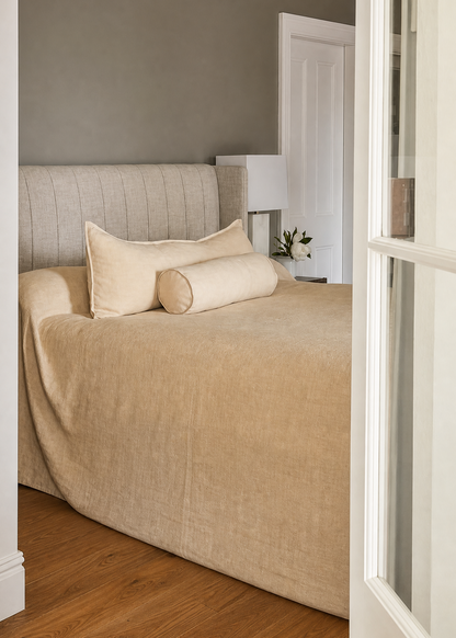 A neatly made bed with a beige textured duvet cover and matching pillows, positioned in a bedroom with a gray wall, wooden floor, and a white door frame on the right.