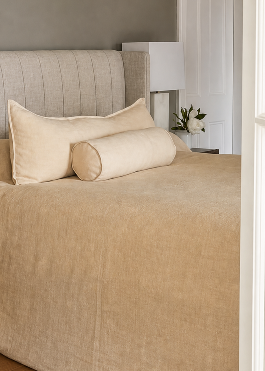 A neatly made bed with a beige textured duvet cover and matching pillows, positioned in a bedroom with a gray wall, wooden floor, and a white door frame on the right.