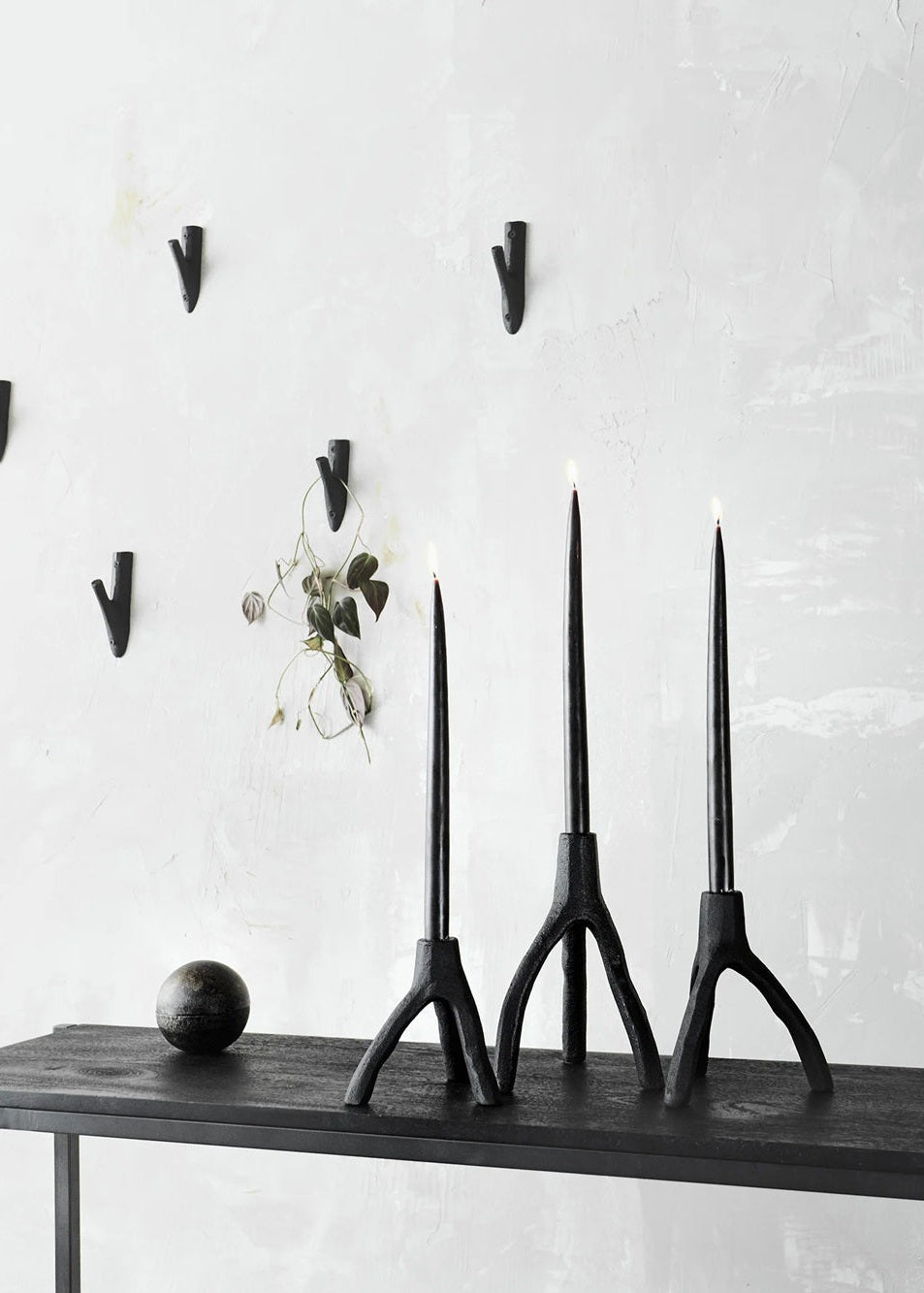 Three black candle holders on a wooden shelf against a white wall with hooks.