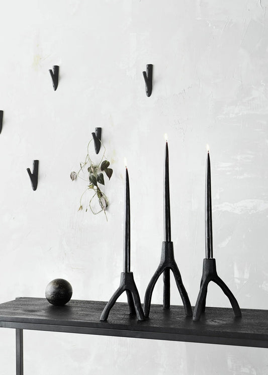 Three black candle holders on a wooden shelf against a white wall with hooks.