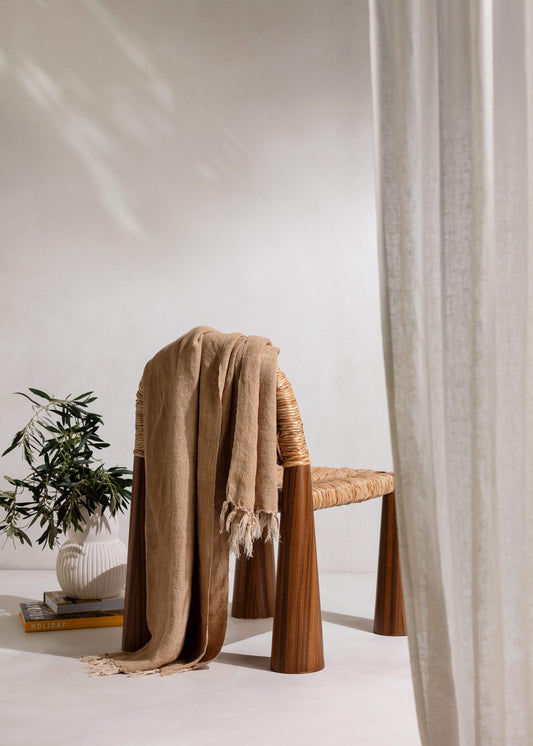 A woven rattan chair with tapered wooden legs draped with a beige fringed throw blanket, styled in a minimalist room with a potted plant and a sheer curtain.